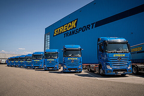 Einige Lastwagen von Streck Transport parken bei Sonnenschein vor der blauen Lagerhalle auf dem Betriebsgelände von Streck Transport in Freiburg