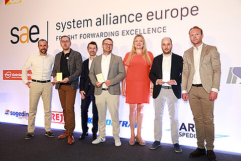 Sieben Personen stehen auf einer Bühne vor einer Wand mit dem Schriftzug „sae | system alliance europe“ und weiteren Logos. Drei Personen halten kleine Auszeichnungen in den Händen, eine Person trägt ein rotes Kleid, die übrigen tragen Anzug oder Business-Kleidung.