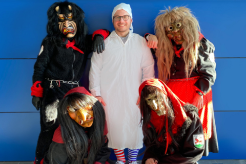 Fünf Personen in traditionellen Fastnachtskostümen und Masken posieren vor einer blauen Wand. In der Mitte steht eine Person in weißem Kostüm mit Haube, Brille und blau-weiß gestreiften Socken, flankiert von vier maskierten Gestalten in schwarzen und roten Gewändern.