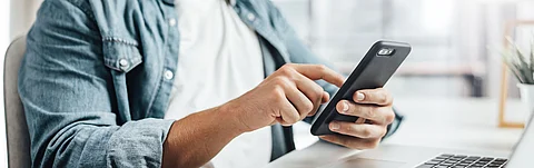 Mann sitzt an einem Tisch mit Notebook und bedient ein Smartphone