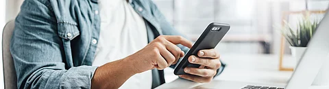 Mann sitzt an einem Tisch mit Notebook und bedient ein Smartphone