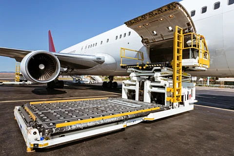 Ein großes Passagierflugzeug steht auf dem Rollfeld mit geöffneter Frachttür. Vor der Öffnung befindet sich eine Hebeplattform mit Rollenbahn und gelben Geländern. Ein Triebwerk und das Fahrwerk des Flugzeugs sind seitlich sichtbar.