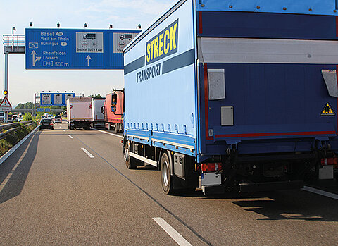 Ein blauer Lastwagen mit der Aufschrift „STRECK TRANSPORT“ fährt auf einer mehrspurigen Autobahn. Über der Fahrbahn befinden sich blaue Wegweiser mit Zielen wie Basel, Weil am Rhein, Huningue, Rheinfelden und Lörrach sowie Hinweisen für den Lkw-Verkehr.
