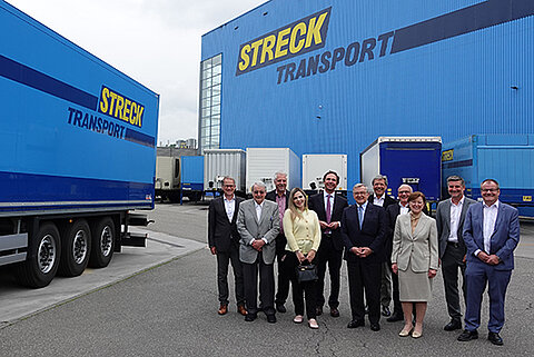 Zehn Personen stehen vor einem blauen Logistikgebäude mit der Aufschrift „STRECK TRANSPORT“. Links im Bild ist ein geparkter Lkw-Anhänger mit demselben Firmenlogo zu sehen.