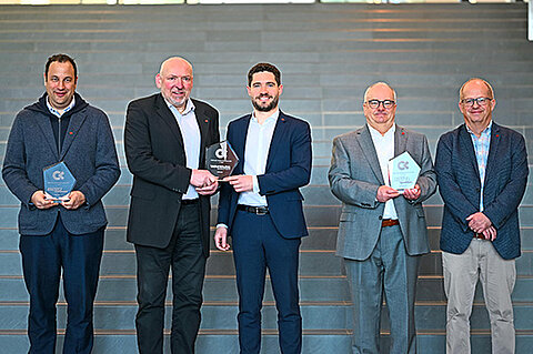 Fünf Personen stehen nebeneinander vor einer breiten Treppe und halten jeweils eine gläserne Auszeichnung in den Händen. Zwei Personen in der Mitte halten gemeinsam eine Trophäe. Alle tragen formelle Kleidung wie Anzüge und Sakkos.