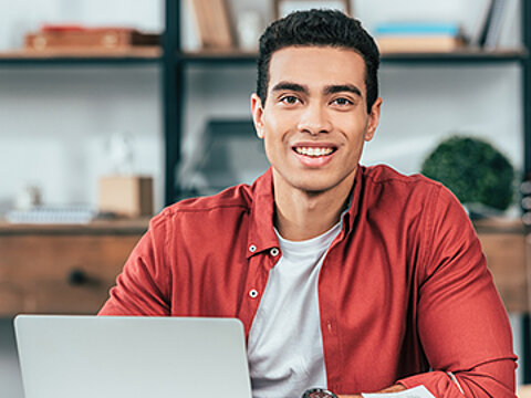Ein Mann Mitte 20 im roten Hemd sitzt mit aufgeschlagenem Buch am Schreibtisch vor einem Notebook