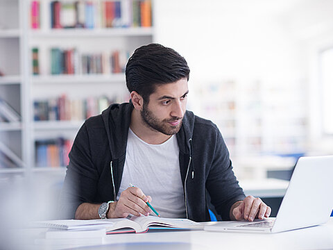 Mann mit Notebook liest konzentriert in einem Buch, im Hintergrund ein Bücherregal