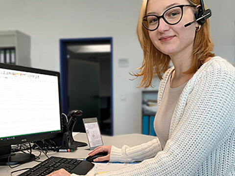 Junge Frau mit Brille und Headset sitzt am Schreibtisch und bedient einen Computer