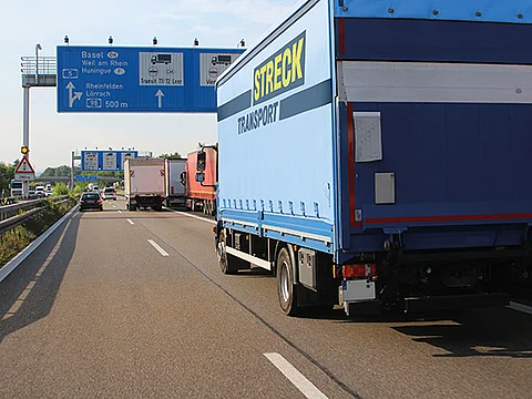 Ein blauer Lkw mit der Aufschrift „Streck Transport“ fährt auf einer mehrspurigen Autobahn. Im Hintergrund sind weitere Lastwagen und Pkw sowie eine blaue Autobahnwegweisung mit Zielen wie Basel, Rheinfelden und Lörrach zu sehen.
