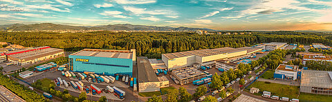 Panorama-Luftbild des Firmengeländes von Streck Transport in Freiburg mit Wechselbrücken und Lkws, im Vordergrund die Blaue Halle und weitere Hallen, im Hintergrund Freiburg und der Schwarzwald