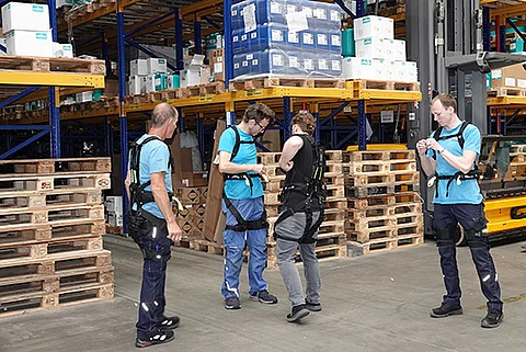 Drei Streck-Mitarbeiter der Kontraktlogistik in Freiburg testen unter Anleitung eines Trainers Exoskelette zur einfacheren und schonenderen Handhabung schwerer Packstücke