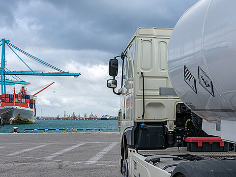 Gefahrgut-Tanklaster in einem Hafen, im Hintergrund ein Containerschiff und zwei Kräne