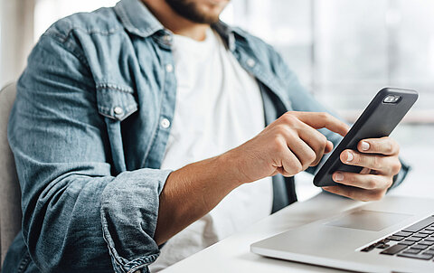 Ansprechpartner Mann sitzt an einem Tisch mit Notebook und bedient ein Smartphone