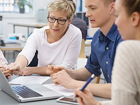 4 Personen sitzen im Büro an einem Tisch und blicken zusammen auf den Bildschirm eines Notebooks