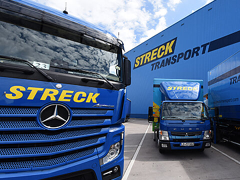 Ein Hybrid-Lkw zwischen zwei Lkws von Streck Transport auf dem Firmengelände in Freiburg, im Hintergrund eine blaue Halle