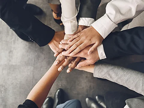 7 Personen legen jeweils eine Hand übereinander, um damit ihre Zusammenarbeit zu zeigen