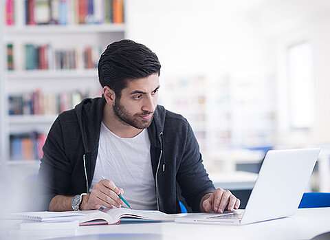 Mann mit Notebook liest konzentriert in einem Buch, im Hintergrund ein Bücherregal