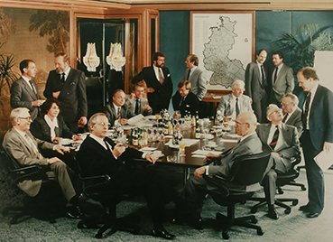 Versammlung der Teilhaber des Deutschen Paketdienstes DPD in den 1980er-Jahren, eine Gruppe von 16 Männern und einer Frau sitzen und stehen um einen großen Tisch mit Unterlagen und Getränken