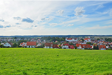 Ansicht der Stadt Meßkirch bei leicht bewölktem Himmel, im Vordergrund eine grüne Wiese, im Hintergrund Gebäude und eine Kirche